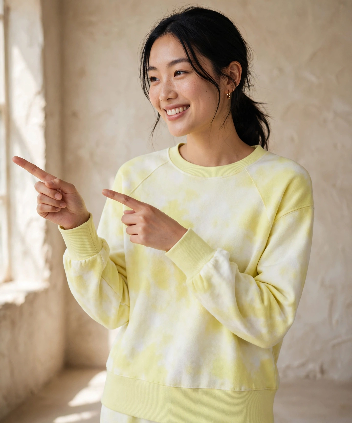 Asian woman with dark hair in ponytail wearing light yellow and white tie-dye crewneck sweatshirt with ribbed cuffs and hem, smiling and pointing both index fingers to the left side, standing in warm beige plaster studio with soft natural window light, portrait 1144 by 1375 pixels, premium editorial, no added text