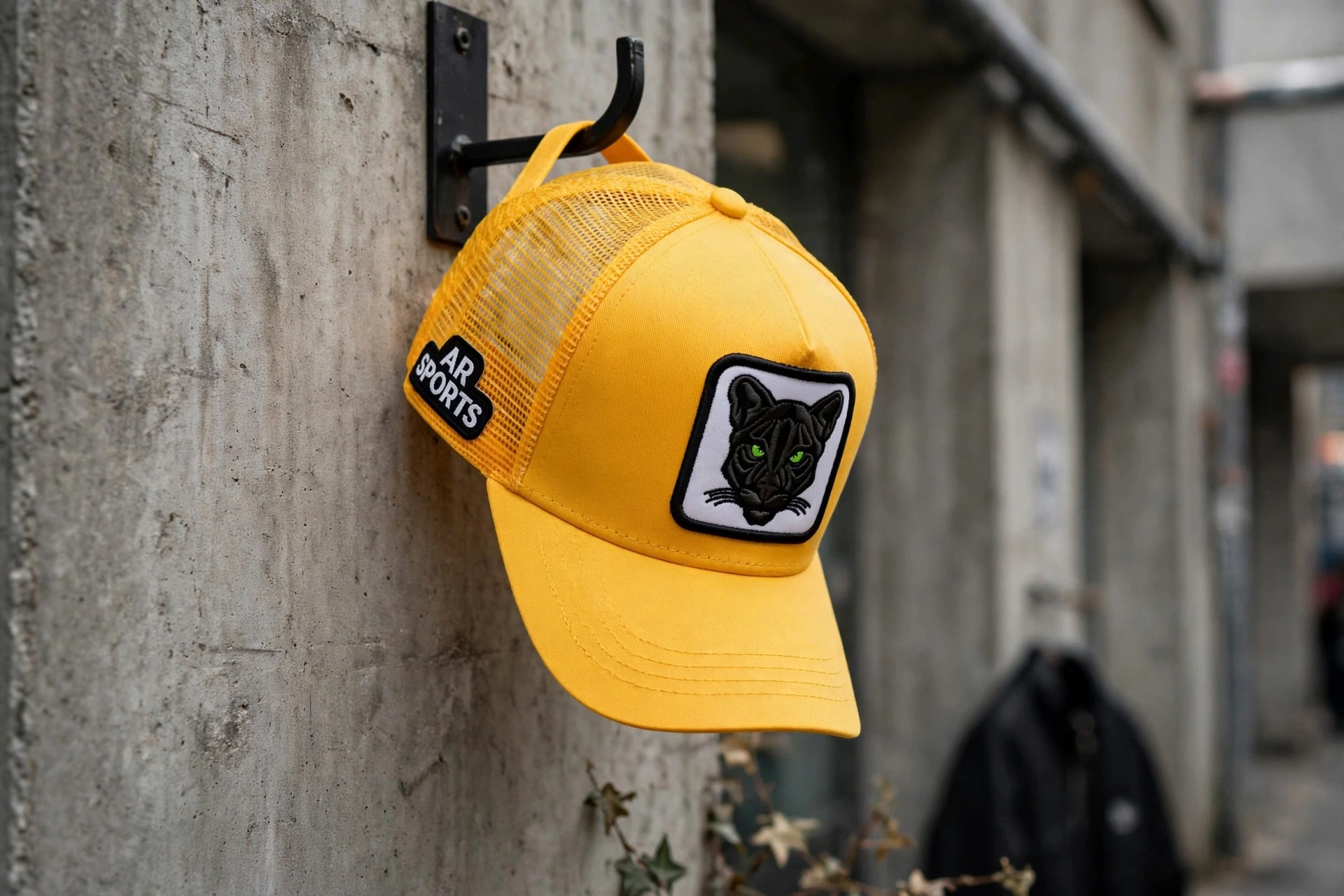 Yellow high-profile trucker cap with black panther patch green eyes and AR SPORTS side patch hanging on concrete wall hook - deep crown flat brim urban streetwear
